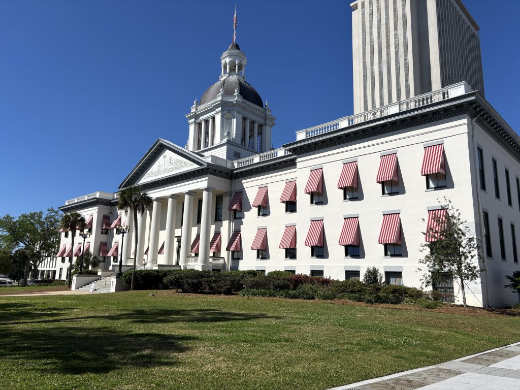 a white building with a dome and red striped awnings over the windows, set against a clear blue sky with a taller structure in the background.