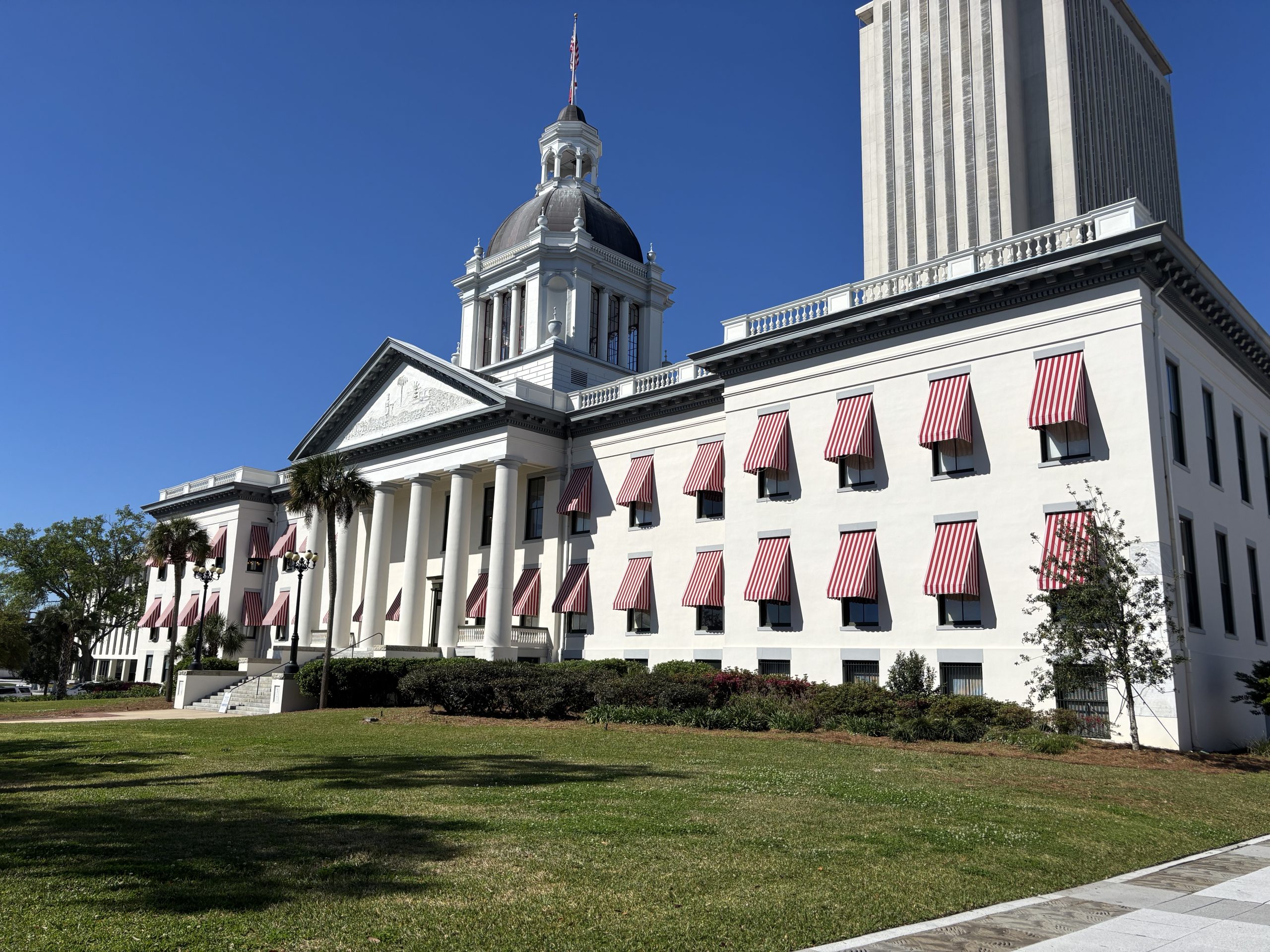 a white building with a dome and red striped awnings over the windows, set against a clear blue sky with a taller structure in the background.