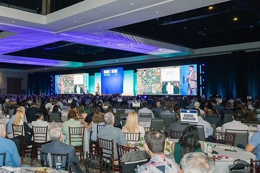 a large audience sits facing a stage with multiple screens displaying maps and a presenter speaking at a conference or seminar.