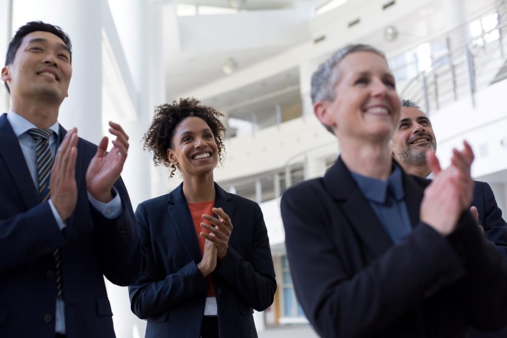 four people in business attire stand indoors, smiling and applauding, with a modern office setting in the background.