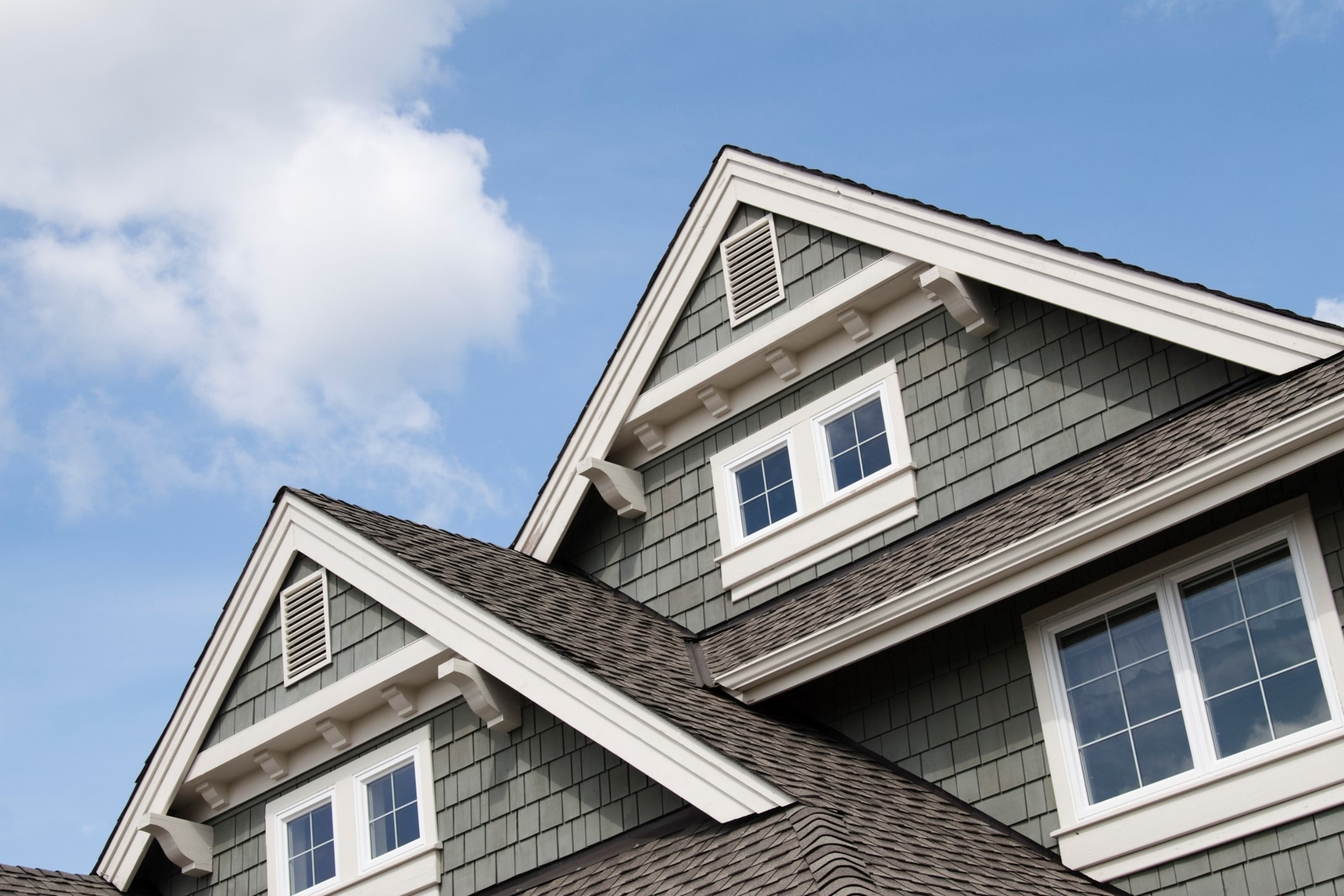 close up of the upper portion of a house with green shingles, white trim, and two gable roofs against a blue sky with scattered clouds.
